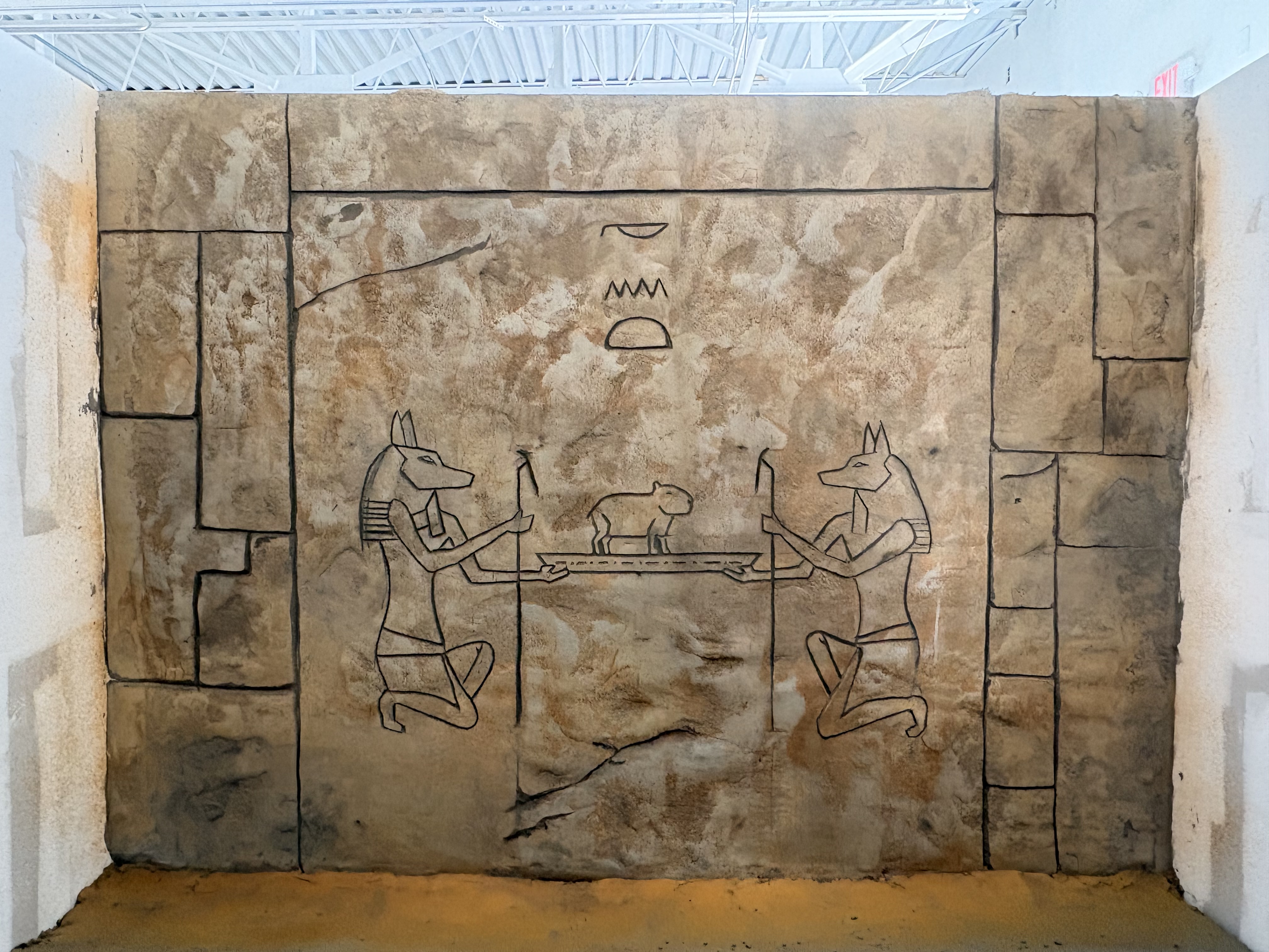 Hand-carved relief scene showing two jackal-headed figures weighing a small capybara on a scale, in an Ancient Egyptian art style